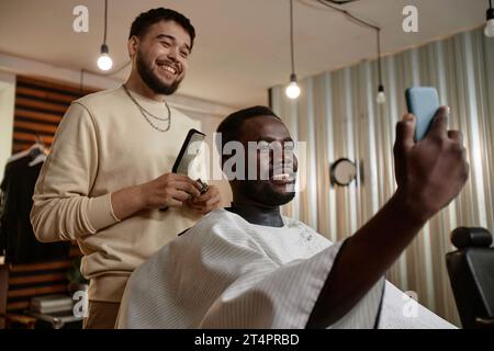 Sorridente uomo afroamericano che fa un ritratto selfie sul suo smartphone mentre il barbiere si taglia i capelli Foto Stock