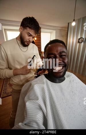 Immagine verticale di un uomo afro-americano che fa un ritratto di selfie mentre si taglia i capelli nel barbiere Foto Stock