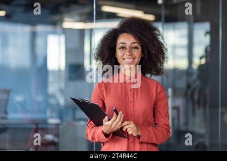 Ritratto di una giovane e bella finanziatrice in camicia rossa all'interno dell'ufficio sul posto di lavoro, donna d'affari sorridente che guarda la macchina fotografica, tiene documenti, cartelle. Foto Stock