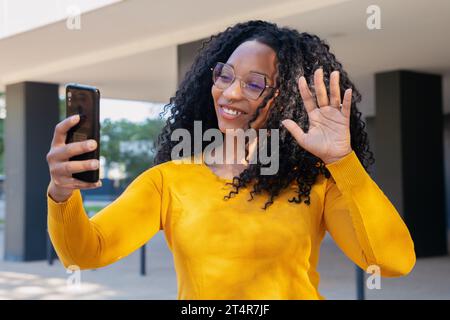 una giovane donna afro-americana sorridente che guarda la fotocamera mentre fa una videoconferenza con il suo smartphone all'aperto. Foto Stock