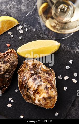 Ostriche fresche chiuse servite con un bicchiere di vino bianco e una fetta di limone su un piatto di ardesia su fondo di marmo nero Foto Stock