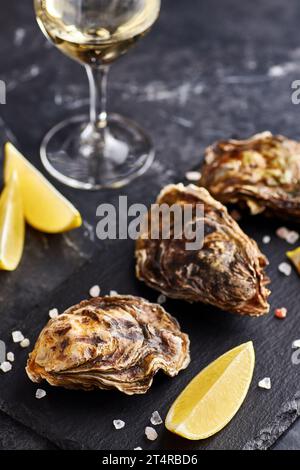 Ostriche fresche chiuse servite con un bicchiere di vino bianco e una fetta di limone su un piatto di ardesia su fondo di marmo nero Foto Stock