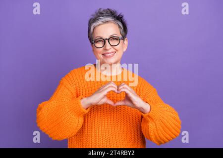 Foto della nonna pensionata che indossa un pullover di tendenza arancione che mostra la società di beneficenza finger Love Heart isolata su sfondo di colore viola Foto Stock