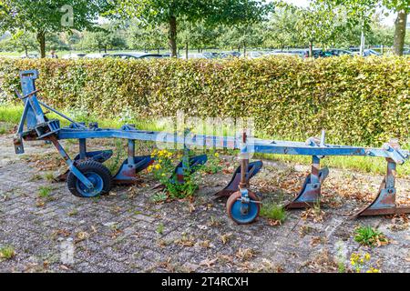 Aratro per trattori agricoli in metallo blu in un'azienda agricola, arrugginito e abbandonato, auto parcheggiate in un'area di parcheggio in sottofondo, cespugli e alberi, giornata di sole a SW Foto Stock