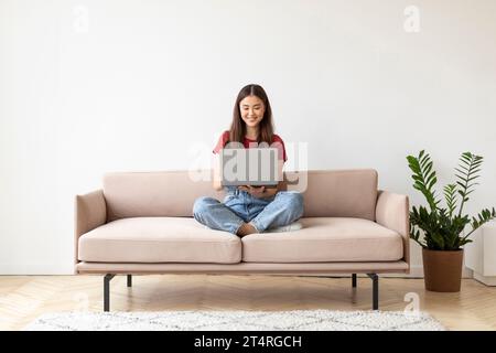 Bella giovane donna asiatica che utilizza il computer portatile mentre si siede sul divano a casa Foto Stock