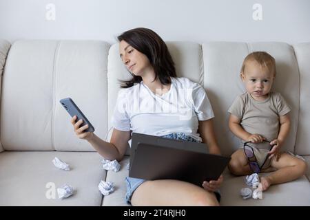 una giovane mamma sta cercando di lavorare a casa con un bambino. Procrastinazione sul lavoro. Foto Stock