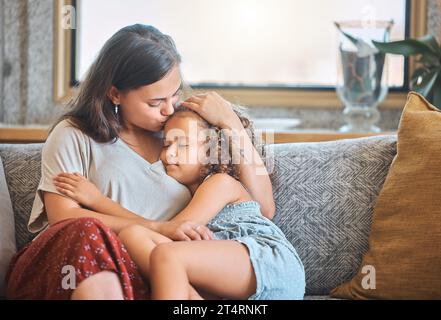 Amata madre che bacia sua figlia sulla fronte mentre si addormenta tra le braccia mentre è seduta sul divano a casa. Bambina stanca che cade Foto Stock