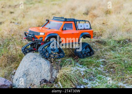 Vista di un veicolo fuoristrada giocattolo radiocomandato arancione con ruote caterpillar su rocce e fondo erboso. Foto Stock