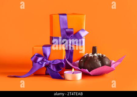 Deliziosa torta al cioccolato, confezioni regalo e candela accesa per festeggiare Halloween su sfondo arancione, primo piano Foto Stock