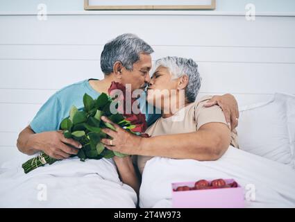 Amore, coppia anziana e bacio con rose a letto, romanticismo e affetto a casa. Fiori, uomo anziano e donna in camera da letto per intimità, cura e.. Foto Stock