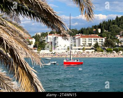 Lo yacht a vela rosso naviga sul mare sullo sfondo di una spiaggia affollata Foto Stock