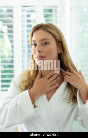 Donna caucasica felice in accappatoio che tocca il collo e sorride nel bagno soleggiato Foto Stock