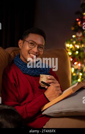 Un uomo asiatico sorridente e felice con una sciarpa e un maglione intimo si rilassa su un divano nel suo soggiorno la notte di Natale, sorseggiando cioccolata calda e leggendo Foto Stock