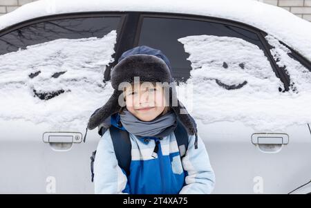 ragazzo con un caldo cappello di pelliccia si trova accanto a un'auto coperta di neve e volti umani dipinti sui finestrini. La creatività dei bambini. Freddo inverno, divertimento W Foto Stock