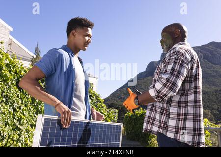 Felice padre afro-americano e figlio adulto con pannello solare che parla nel giardino soleggiato Foto Stock