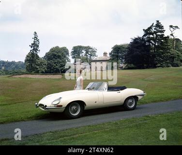 Foto pubblicitaria iniziale di Jaguar e-TYPE S1, Open a due posti, 3,8L, con modello in abito bianco all'Halesowen Golf Club con clubhouse sullo sfondo. Foto Stock