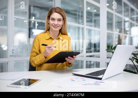 Ritratto donna di successo felice finanziatrice che fa documenti, donna d'affari che tiene in mano documenti, documenti, contratti e rapporti, sorride con gioia, è soddisfatta di lavorare con il notebook, usando la calcolatrice Foto Stock