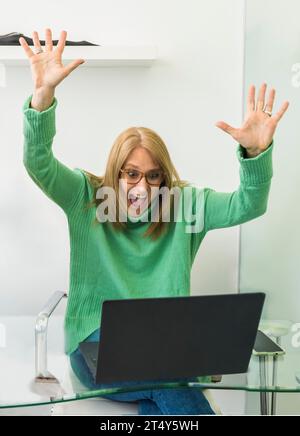 Ritratto di una donna adulta entusiasta che festeggia il successo, alzando le braccia alzando la mano mentre usi un computer portatile. Concetto Win Foto Stock
