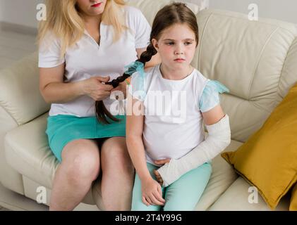 Adorabile bambina prescolare con un braccio rotto, fusa ortopedica a casa sul divano con la madre. Arto rotto e braccio rotto in un gesso nei bambini Foto Stock