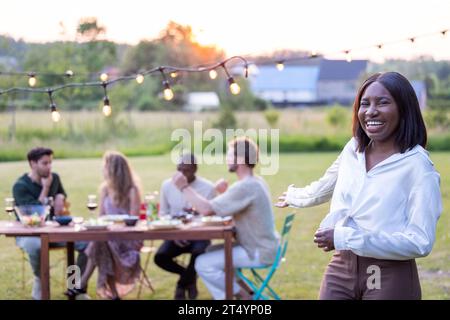 Un incontro gioioso in una giornata di sole racchiude una bella giovane donna nera in una camicetta bianca, ridendo e assaporando un barbecue in giardino tra amici, vino e divertimento al picnic all'aperto. L'essenza del cameratismo e dei festeggiamenti estivi è catturata in modo impeccabile ad alta risoluzione. Giovane donna radiosa che si gode il barbecue Sunny Garden con gli amici. Foto di alta qualità Foto Stock