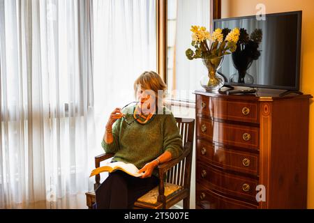 Donna matura seduta accanto alla luce della finestra che si diverte a leggere Foto Stock