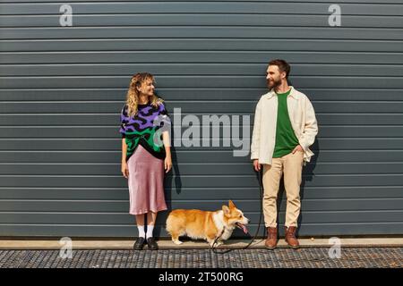 coppia felice ed elegante che cammina con il cane corgi vicino alla porta grigia del garage, compagni di animali Foto Stock