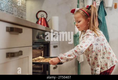 Piccola ragazza carina che fa biscotti dolci fatti in casa. Ha messo una teglia nel forno caldo. Preparazione dei biscotti di Natale. Casa dolce casa e caldo f Foto Stock