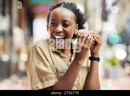 Donna nera e felice che mangia un panino, un pranzo o un cibo in città, un ristorante urbano o un cliente in pausa dal lavoro con felicità, hamburger, pasti o. Foto Stock