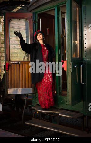 Attraente giovane donna in abito flapper rosso 1920s e cappello di cloche che viaggia con un antico treno a vapore Foto Stock