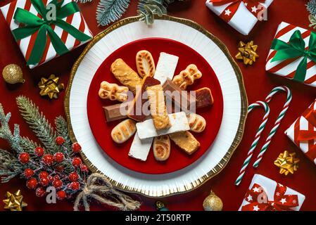 Vista dall'alto del torrone di natale dolce con ornamenti natalizi su un piatto su sfondo rosso. Assortimento di dolci natalizi tipici della Spagna Foto Stock