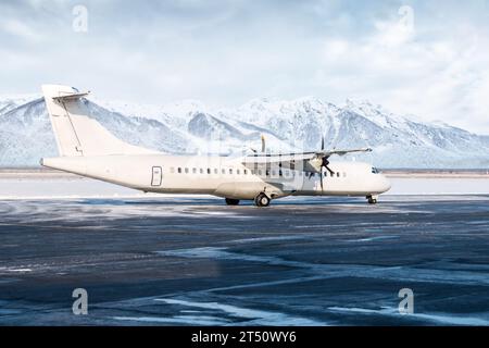 Aereo di linea a turboelica per passeggeri bianco sul grembiule invernale dell'aeroporto sullo sfondo di alte e pittoresche montagne Foto Stock