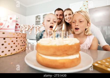 Compleanno, torta e festeggiamenti con una ragazza e sua sorella che soffiano le candele e fanno un desiderio con la sua famiglia sullo sfondo. Festeggiamo, festa Foto Stock