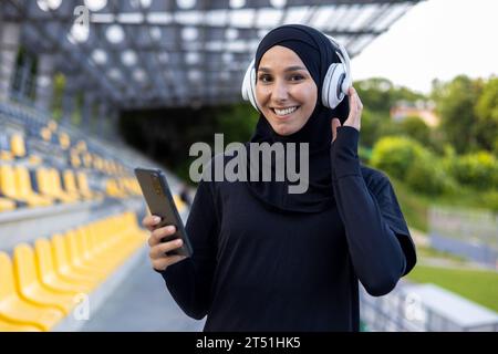 Ritratto di una giovane donna musulmana hijab in uno stadio sportivo che corre e fa esercizi fisici attivi, sorride e guarda la fotocamera, utilizzando il telefono e le cuffie per ascoltare musica. Foto Stock