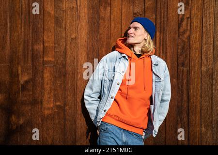 Sorridente biondo Guy aspetto scandinavo androgino godendosi il sole in piedi vicino alla recinzione di legno. Foto Stock