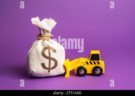 Un bulldozer spinge un sacco di soldi da un dollaro. Uso inefficace dei fondi. Finanziamento dei lavori di smantellamento. Servizi di demolizione, livellamento del terreno. Lavoratore industriale Foto Stock