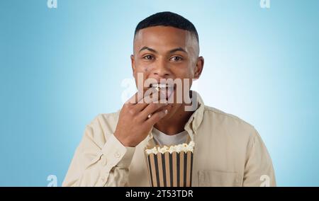 Ritratto, uomo felice e popcorn per il cinema in studio su sfondo blu per il mockup dell'intrattenimento. Modello maschile, mangiare e affamato con delizioso spuntino Foto Stock