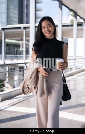 Un ritratto di una donna d'affari asiatica millenaria, bella e sicura di sé, tiene in mano il suo vestito e una tazza di caffè da asporto mentre cammina sullo Skywalk Foto Stock