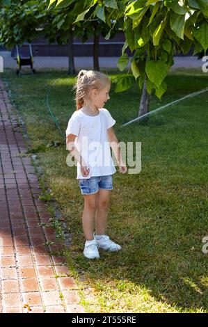 Bambina che cammina sott'acqua dall'irrigazione sprinkler nel parco cittadino estivo. Concetto di infanzia, tempo libero e persone - Happy Children Rest an Foto Stock