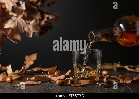 Il whisky viene versato da una bottiglia in un bicchiere. Whiskey su un tavolo nero con foglie di quercia essiccate. Foto Stock