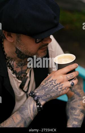 L'uomo bello ed elegante beve caffè, le sue mani sono coperte di tatuaggi. Foto Stock