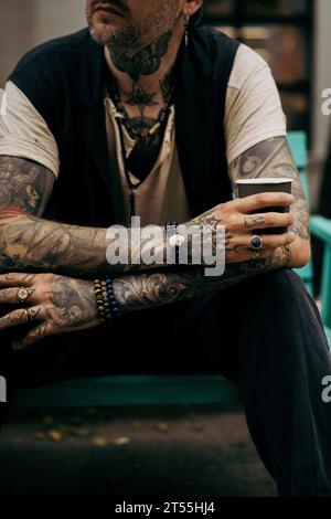 L'uomo bello ed elegante beve caffè, le sue mani sono coperte di tatuaggi. Foto Stock