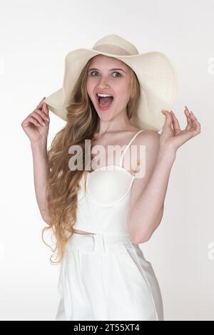 Espressione facciale di una donna bionda che guarda la macchina fotografica, braccia sollevate e un cappello di paglia floppy con le mani. Modello di felicità ritratto Foto Stock