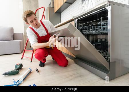 Il tecnico o il lavoratore in uniforme installa la lavastoviglie nei mobili della cucina. Il riparatore indossa tuta da lavoro riparando la manutenzione della lavastoviglie. Maestro Foto Stock