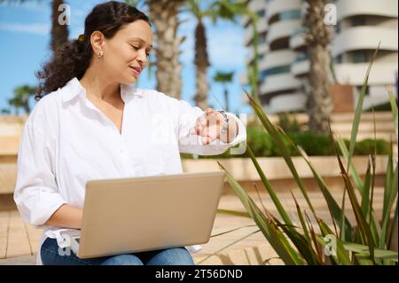 Donna d'affari sorridente con camicia casual bianca, che controlla il tempo sul suo orologio da polso, lavora online su un notebook contro un grattacielo Foto Stock