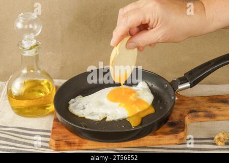 Donna a mano immergendo il pane sul tuorlo di un uovo fritto in olio d'oliva con una padella su una tavola di legno Foto Stock