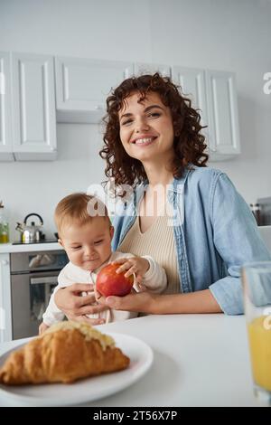 mamma allegra che guarda la macchina fotografica durante la colazione con la bambina vicino alla mela matura e al gustoso croissant Foto Stock