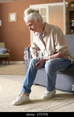 Immagine verticale di una donna anziana seduta su un divano in salotto e affetta da dolori alle gambe Foto Stock