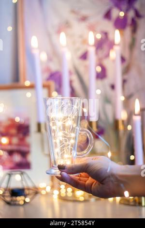 Mano di una donna che tiene una tazza di caffè in vetro con luci a corda. Bokeh, candele in fiamme, atmosfera natalizia invernale. Immagini calde e stimolanti Foto Stock