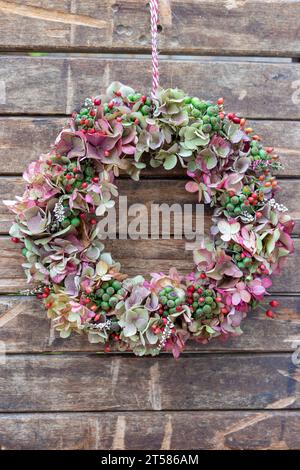 corona di ortensie, fianchi di rosa e bacche di edera appese al muro di legno Foto Stock