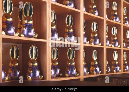 Esposizione di coniglietti pasquali al cioccolato, ciascuno avvolto in lamina d'oro e argento decorata. Sono allineate in modo ordinato su ripiani in legno rustico, che mostrano il de Foto Stock